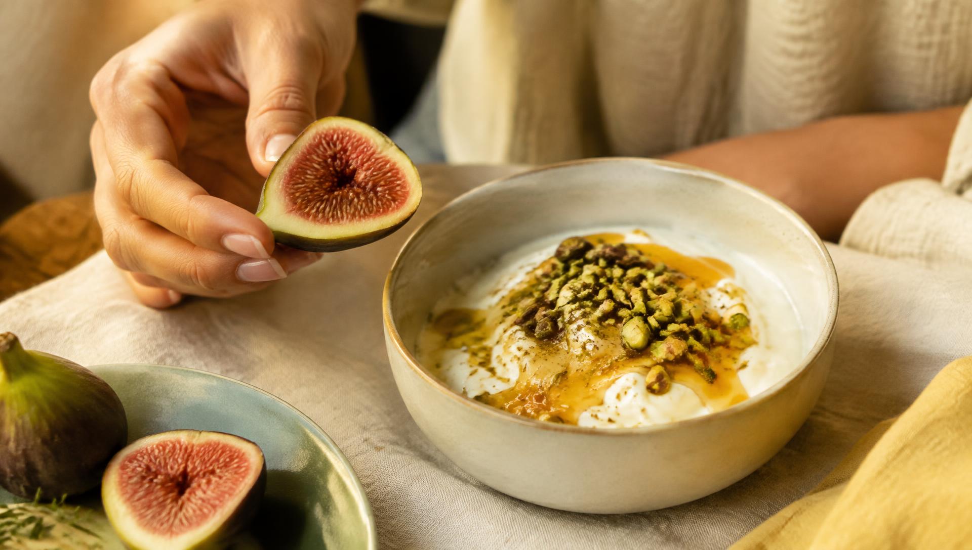 Hands holding a fresh fig beside a bowl of yoghurt
